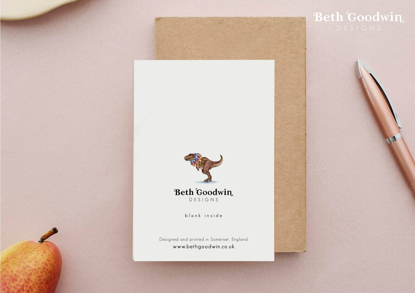 Notebook with a dinosaur illustration and 'Beth Goodwin Designs' branding on a pink surface.