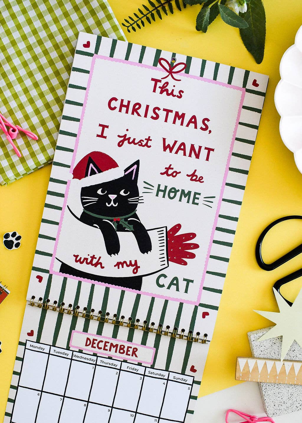 A Christmas-themed wall calendar page with a black cat wearing a Santa hat and scarf, surrounded by holiday decorations on a bright yellow surface.