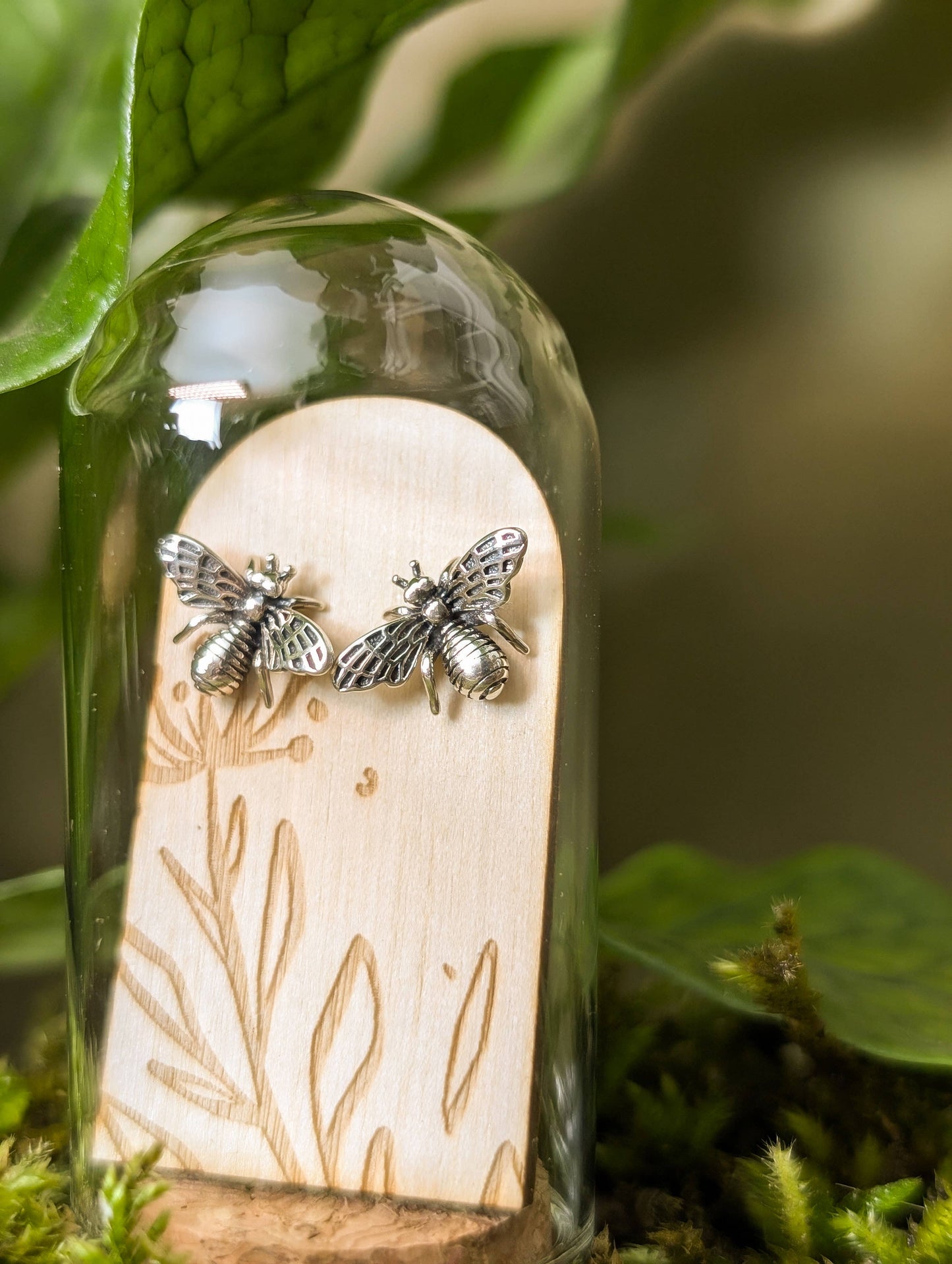 Silver bee-shaped earrings inside a glass dome with a wooden base.