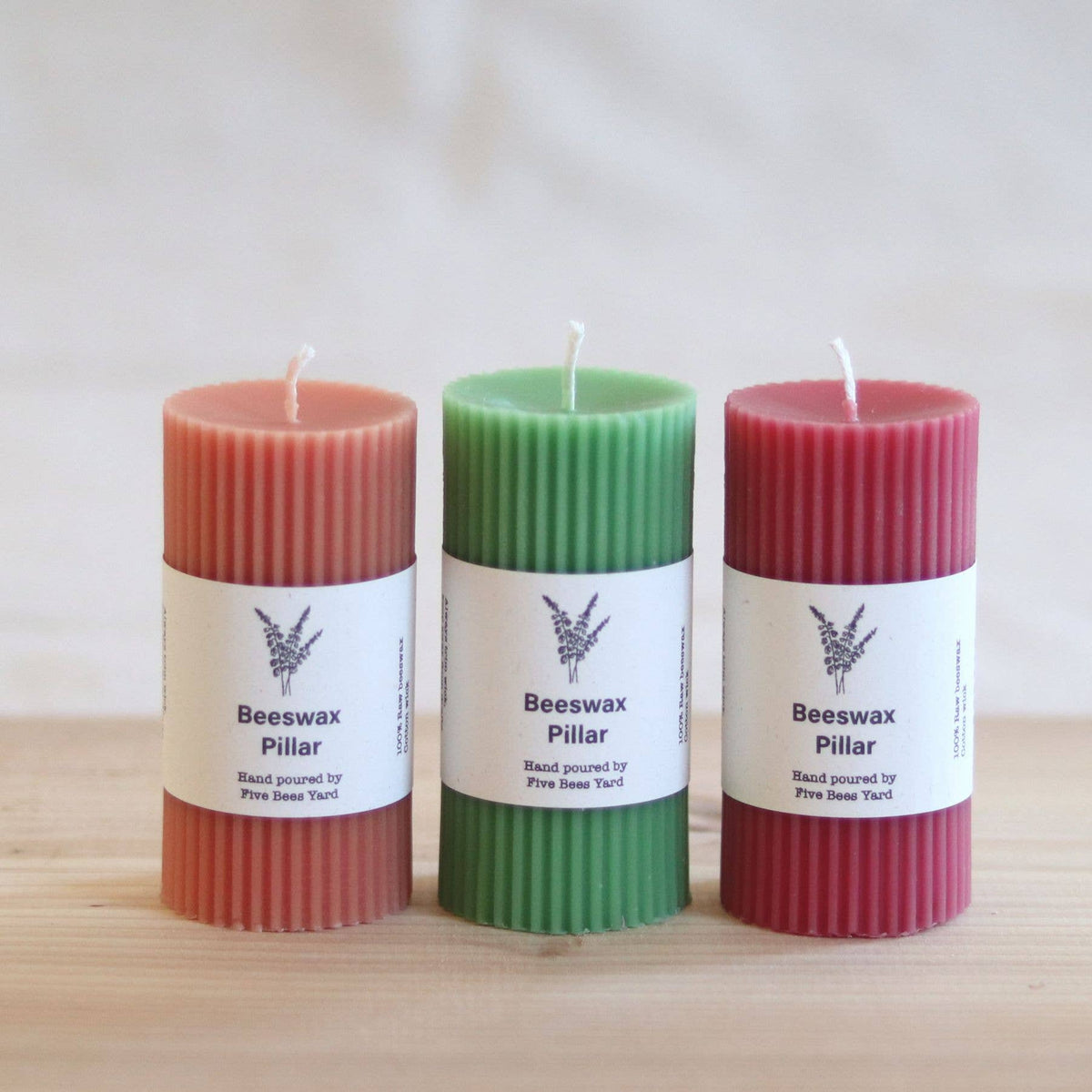 Three hand-poured beeswax pillar candles in coral pink, green, and burgundy red, standing side-by-side on a light wood surface against a soft, neutral background. Each candle features a white paper label with the text 'Beeswax Pillar' and 'Hand poured by Pine Tree Yard'.