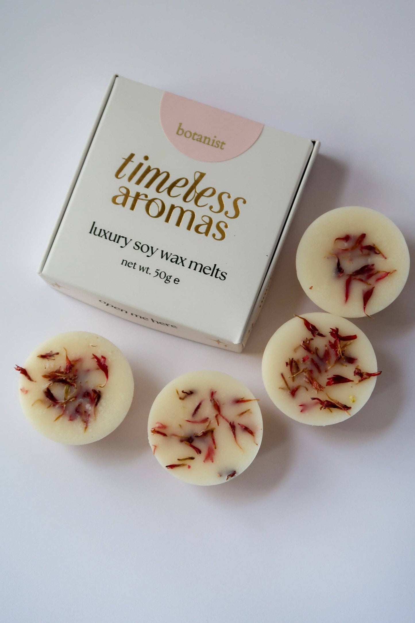Four round cream-colored soy wax melts with red botanical flecks, arranged next to their white and gold Botanist 'Timeless Aromas' packaging box on a clean white background.