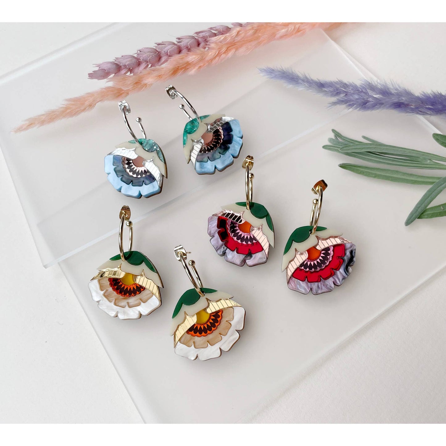 Five pairs of handmade flower-shaped earrings with gold-tone hooks and colorful enamel details, arranged on a white surface with dried decorative grasses.