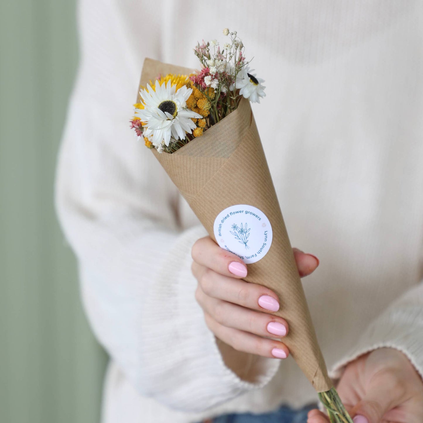 A person in a white knit sweater holds a small bouquet of dried flowers wrapped in brown kraft paper with a round white sticker. The flowers include white, yellow, and pink blossoms.