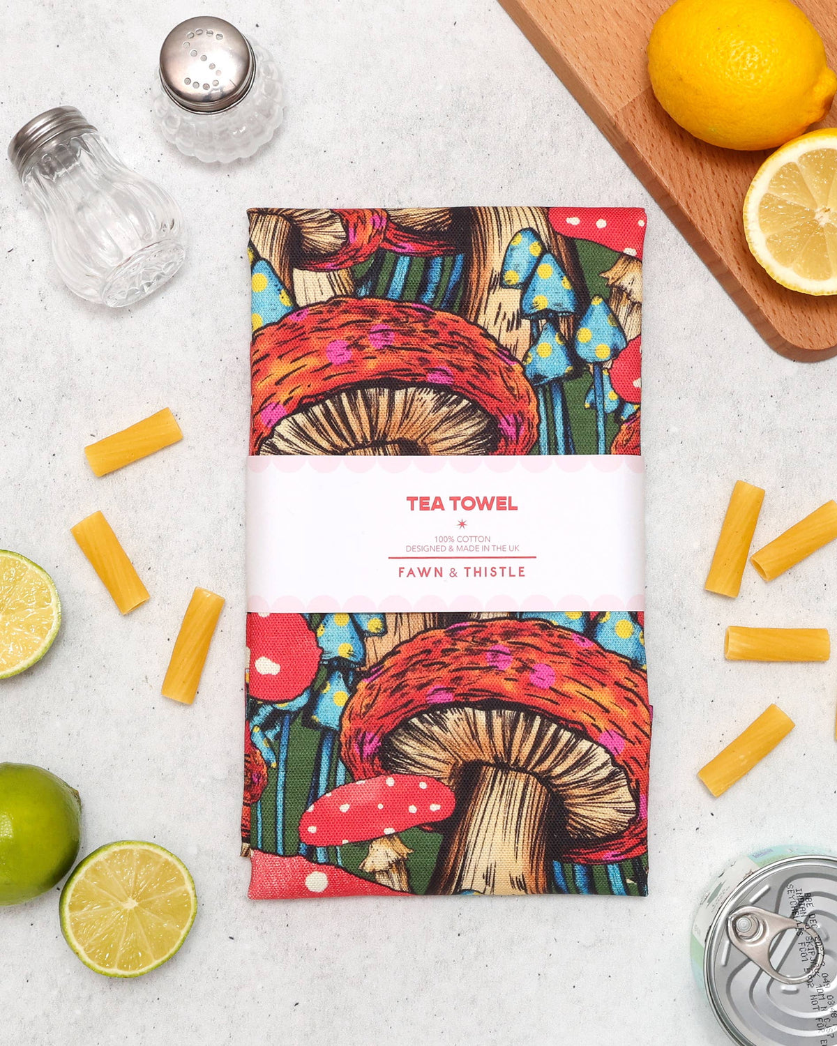 A patterned tea towel with a colorful mushroom illustration and a white paper band reading 'TEA TOWEL' and 'FAWN & THISTLE', arranged on a light surface with lemons, cheese sticks, salt and pepper shakers, and a can around it.