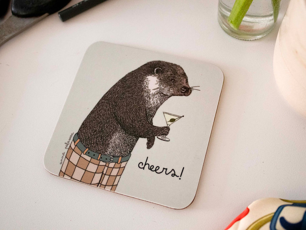 A square white coaster with rounded corners featuring an illustration of an otter holding a martini glass and wearing checked trousers, with the word 'cheers!' written beside them.