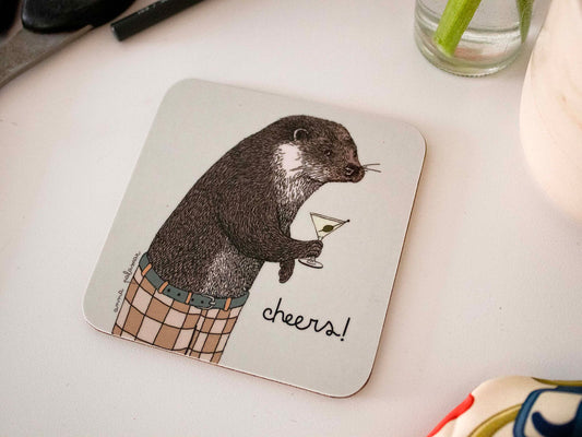 A square white coaster with rounded corners featuring an illustration of an otter holding a martini glass and wearing checked trousers, with the word 'cheers!' written beside them.