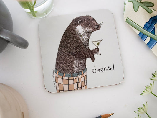 A square white coaster with rounded corners featuring an illustration of an otter holding a martini glass and wearing checked trousers, with the word 'cheers!' written beside them.