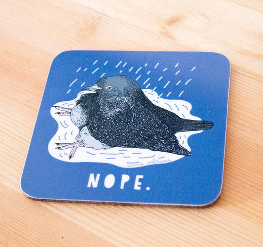 A square blue coaster with rounded corners featuring an illustration of a pigeon in the rain with the word 'NOPE.' printed below, placed on a light wood surface.
