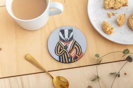 A round coaster featuring an illustration of a badger in a striped jacket and red bow tie, enjoying a little biscuit.