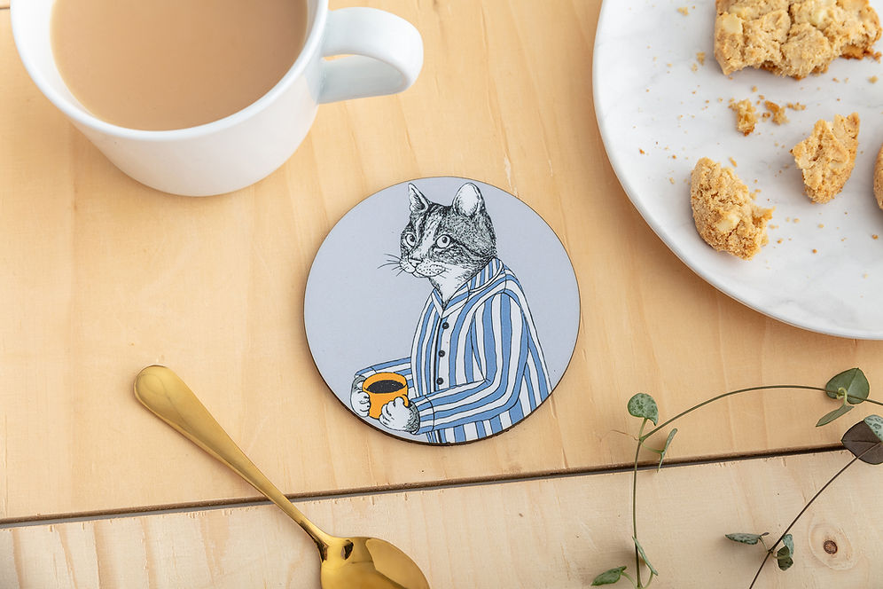 A round coaster featuring an illustration of a cat in blue striped pyjamas holding a mug of coffee.