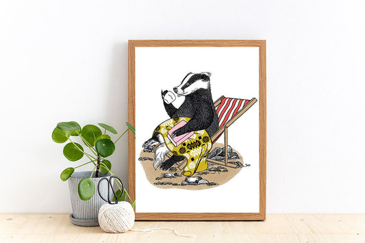 A framed art print featuring a badger relaxing with a cosy yellow blanket on his legs, sipping from a 'cariad' mug on his red and white striped deck chair.