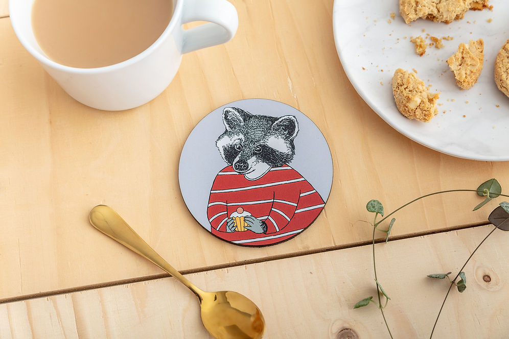 A round coaster featuring an illustration of a raccoon in a red striped top, about to enjoy a little cake.