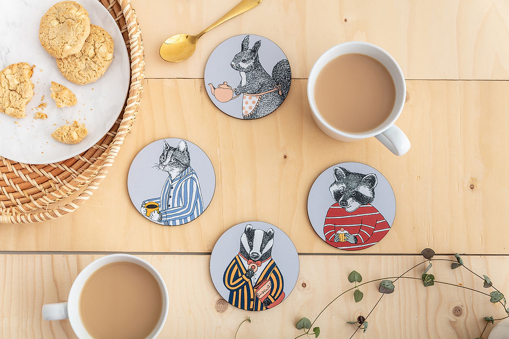 Four round coasters with whimsical animal illustrations on a light wood tabletop. The coasters feature a grey squirrel in a little apron holding a pink teapot, a cat in blue pyjamas, a raccoon in a red striped top, and a badger in a striped blazer.