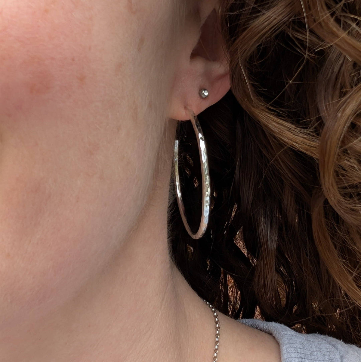 A close-up of a person's ear wearing a large silver hoop earring and a small silver stud earring, with brown hair and part of the neck visible.
