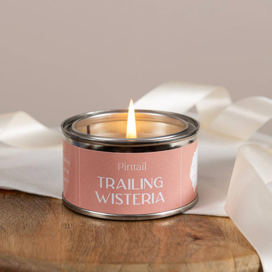 Candle in a tin labeled 'Trailing Wisteria' by Pintail on a wooden surface with white fabric.