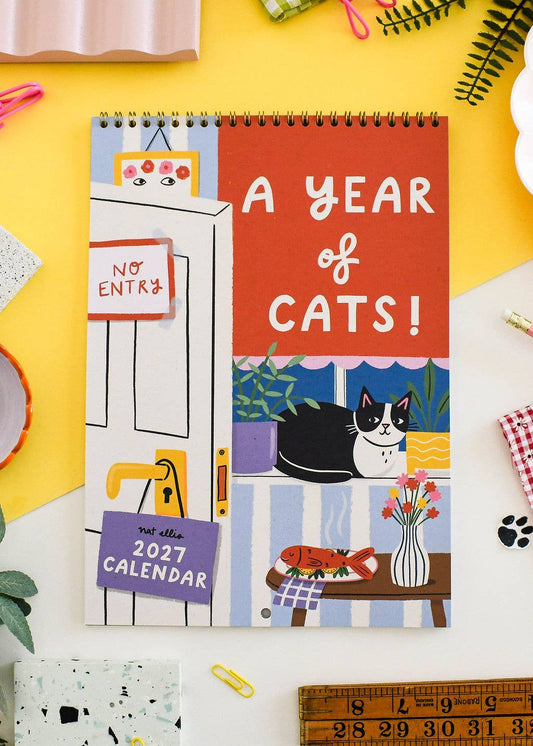 A spiral-bound 2027 calendar titled 'A YEAR of CATS!' laid flat on a bright yellow surface. The cover features colorful cat-themed illustrations, including a black-and-white cat, a fish, flowers, and a door with a 'NO ENTRY' sign.