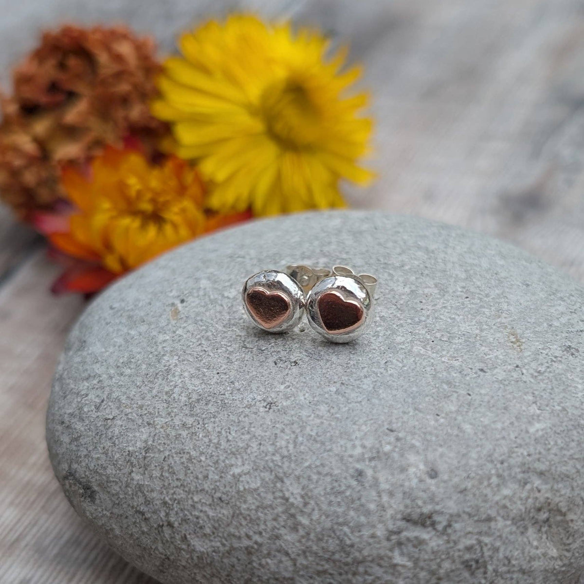 A pair of silver heart stud earrings with dark red heart inlays, displayed on a smooth gray stone with blurred yellow and orange flowers in the background.