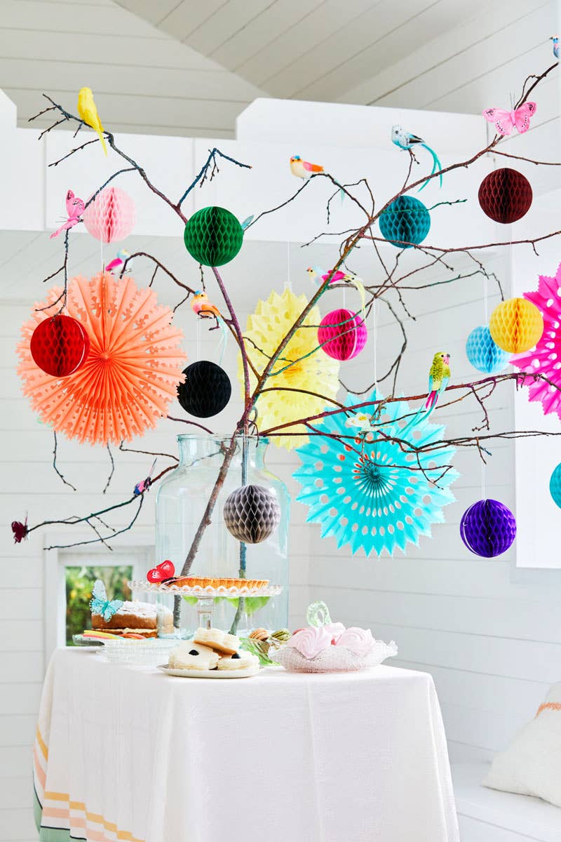 Decorative setup with colorful paper fans and pom-poms on a branch against a white wall.