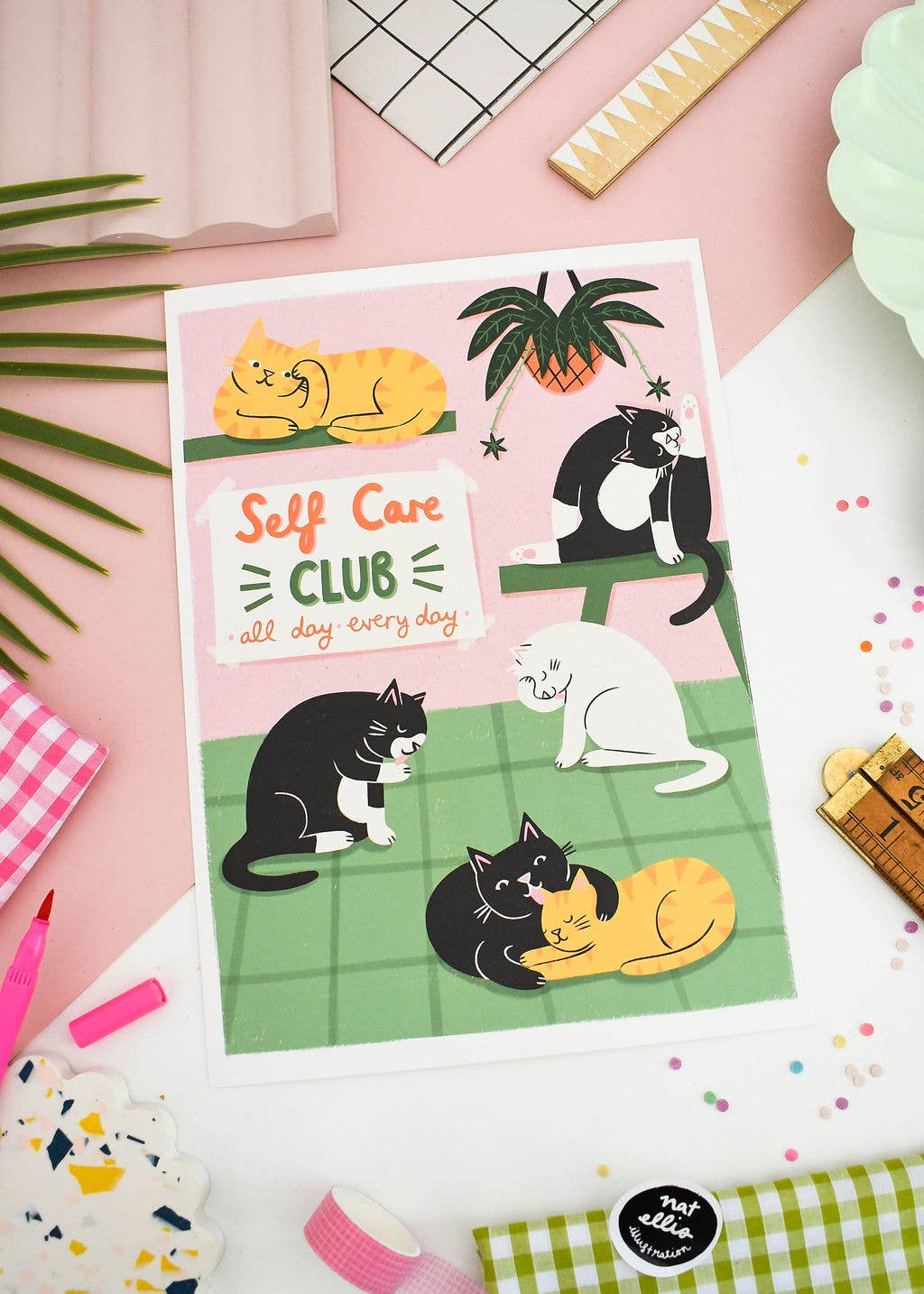 Poster with cartoon cats and 'Self Care Club' text on a pink surface with stationery items.