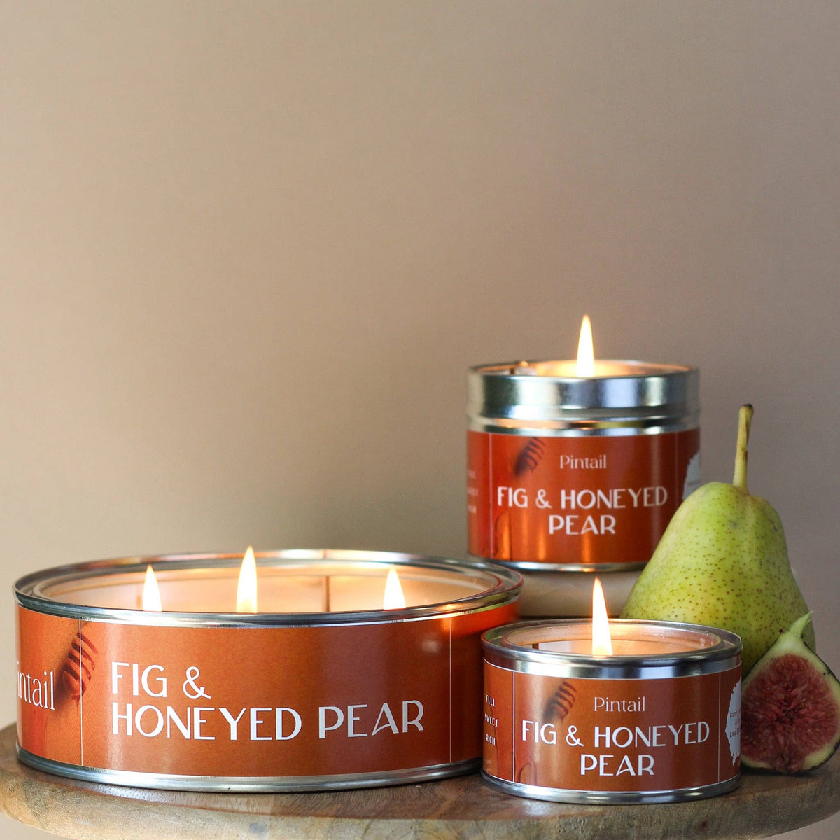 Three 'Fig & Honeyed Pear' candles in tins with a pear and fig on a wooden surface.