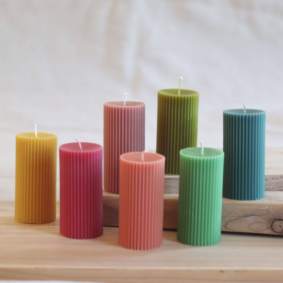 Six ribbed pillar candles in assorted pastel and earthy colors, including yellow, pink, mauve, olive green, coral pink, mint green, and teal blue, arranged on a wooden block over a light wood surface.