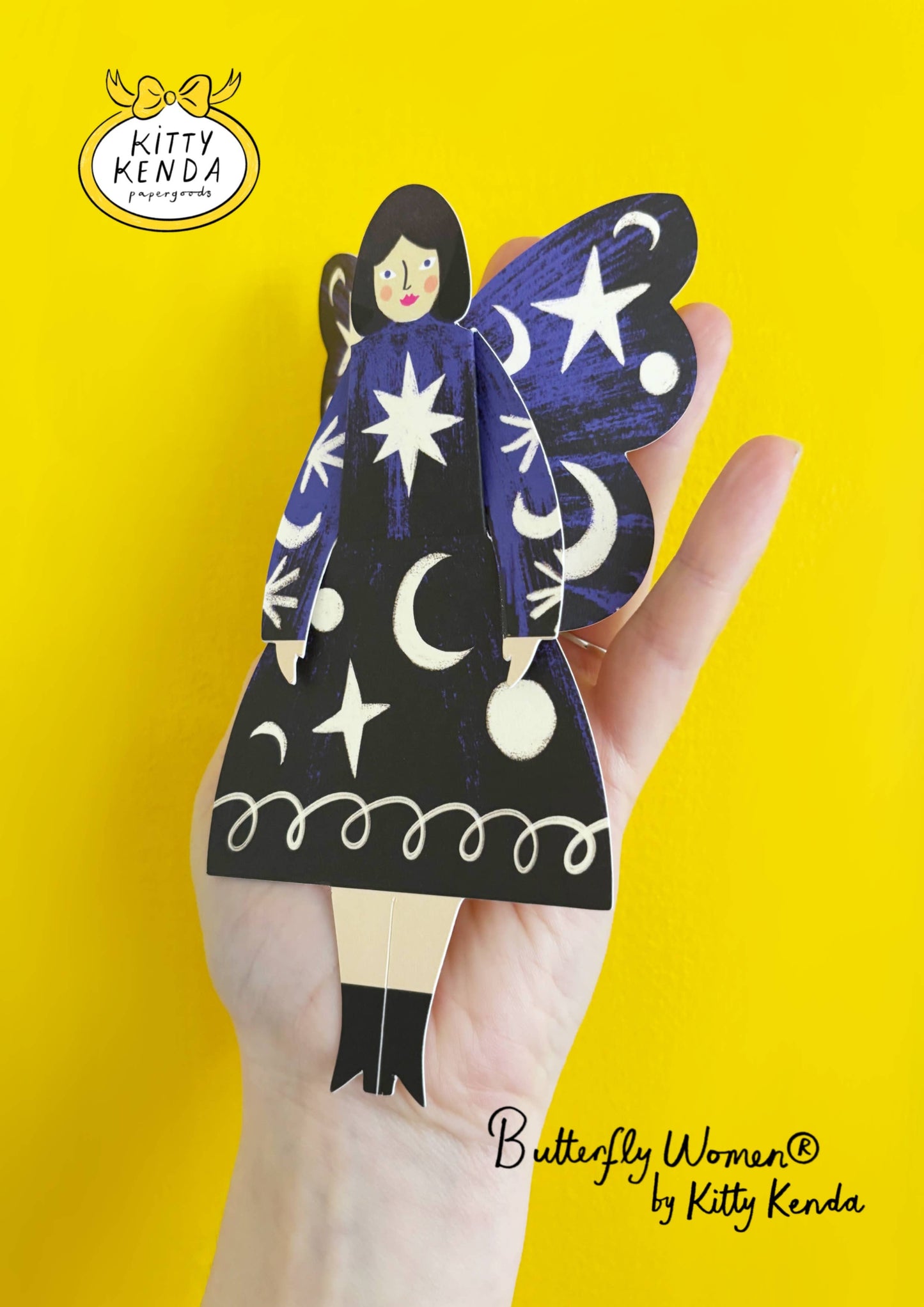 Butterfly-shaped product with star and moon designs held by a hand against a yellow background, branded 'Kitty Kenda'.