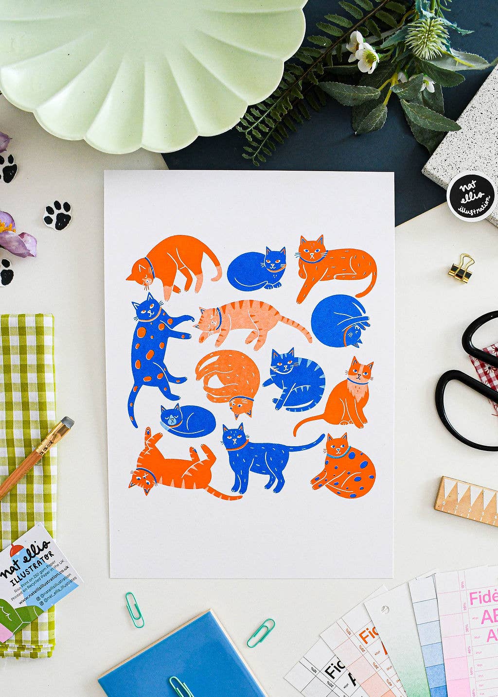 A white art print featuring colorful illustrations of various cats in orange and blue, styled in a flat-lay desk setting with notebooks, scissors, and a mint green dish.