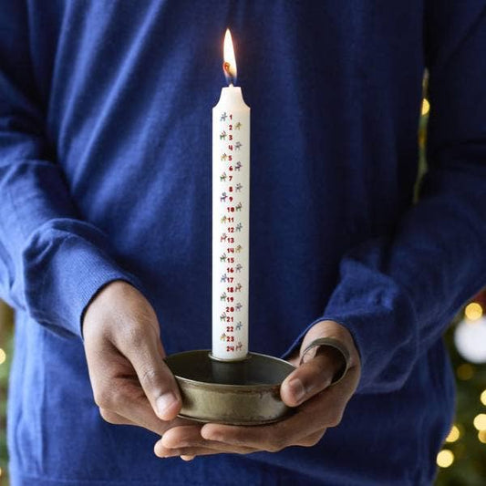 Person holding a lit candle with Christmas designs in a small metal holder against a blurred festive background.