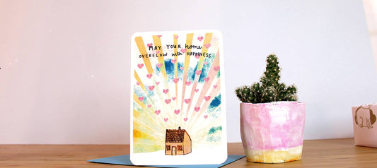 New Home Card, Image of a little house with rays and hearts emanating from it with the words May your home overflow with happiness.