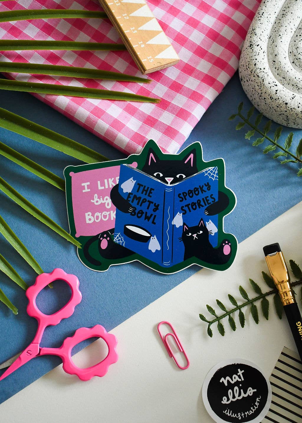 A die-cut sticker featuring a cartoon black cat reading two books, one pink and one blue, placed on a styled flat-lay desk with pink scissors, a pink gingham cloth, green leaves, and stationery.