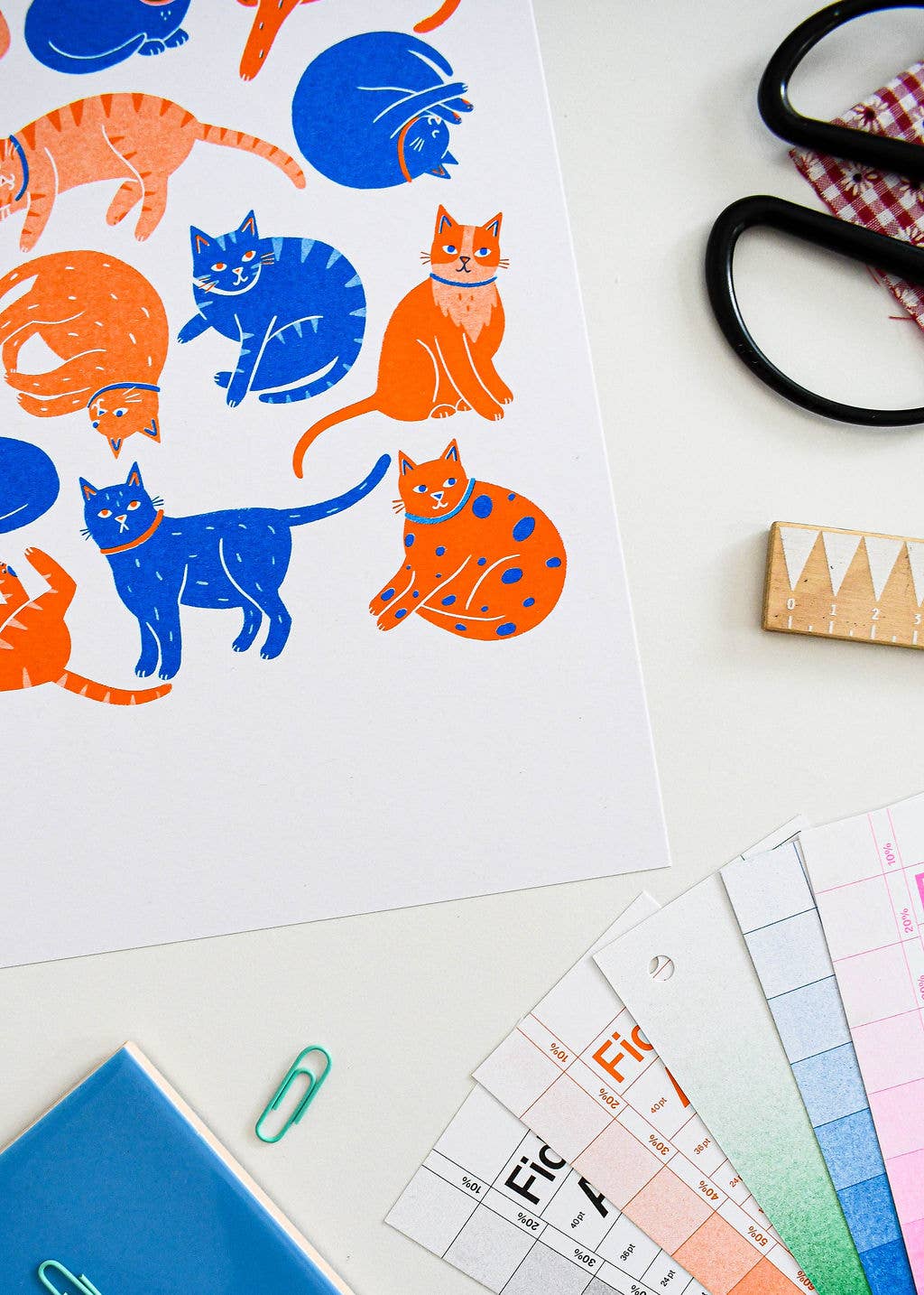 A white art print featuring colorful illustrations of cats in blue, orange, and navy, styled on a white desk with scissors, a wooden stamp, paper clips, and color swatch cards.