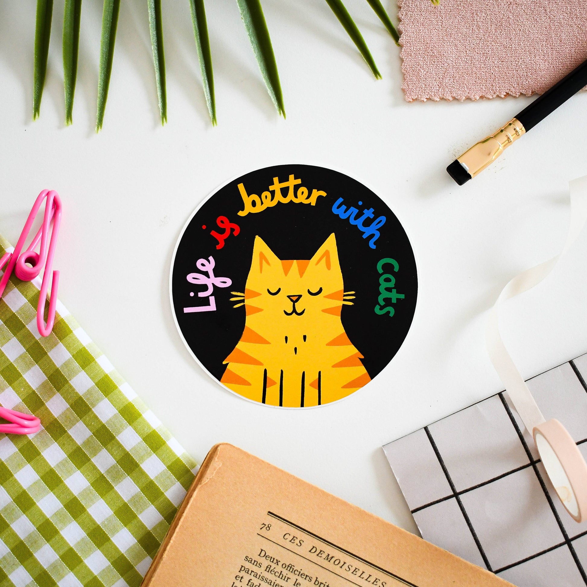 Circular sticker with a cat illustration and text 'Life is better with cats' on a desk with stationery items.