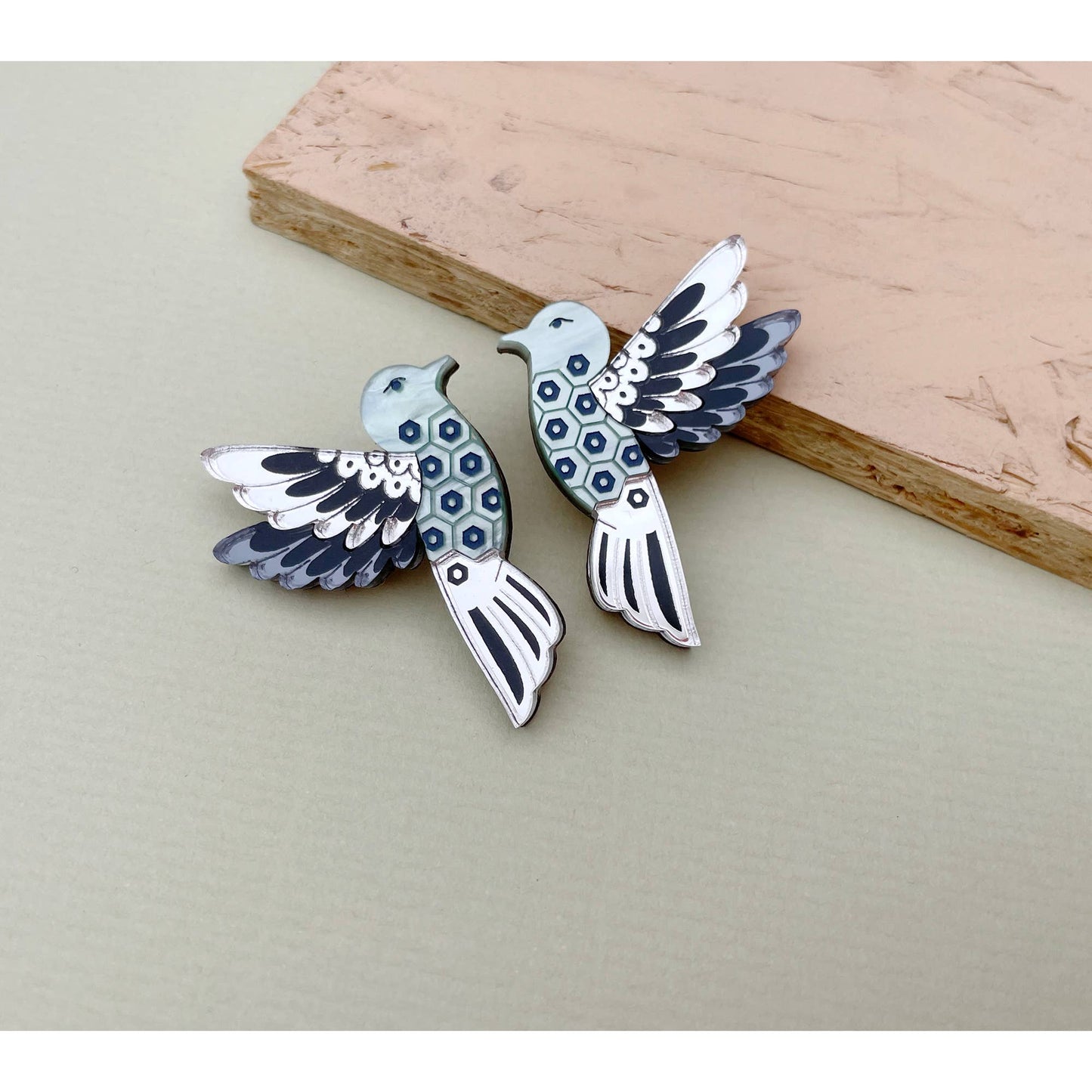 A pair of silver-toned bird earrings with blue and black enamel details, displayed on a neutral beige surface with a wooden block prop.