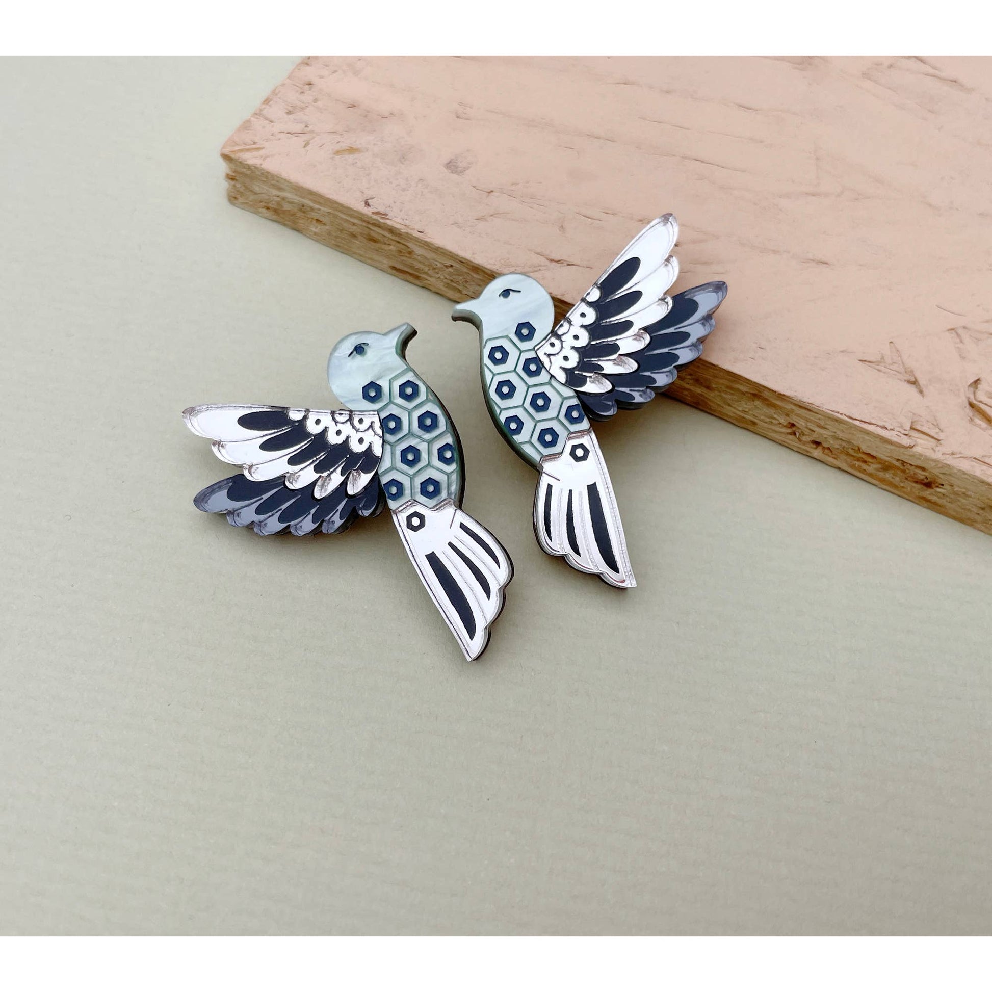 A pair of silver-toned bird earrings with blue and black enamel details, displayed on a neutral beige surface with a wooden block prop.
