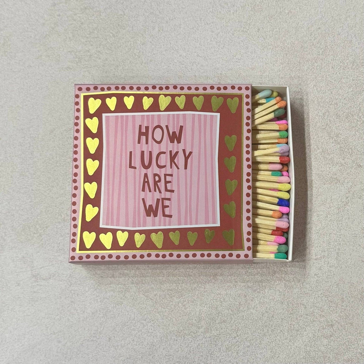 Matchbox with 'How Lucky Are We' text and colorful matches on a light gray background
