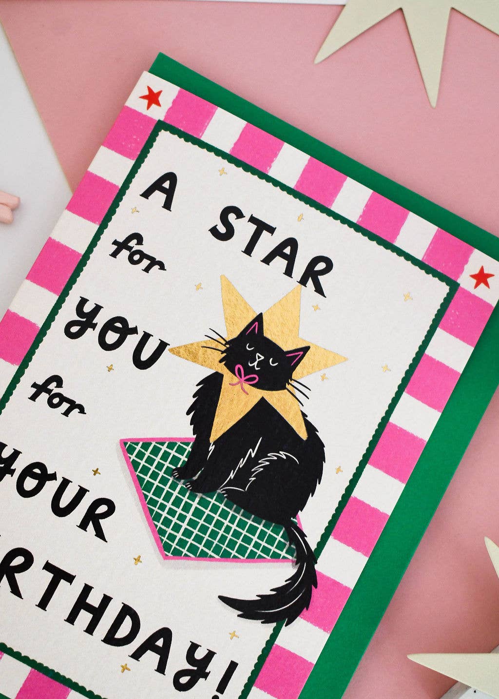 A handmade-style birthday card featuring a black cat wearing a yellow star collar, with the text 'A STAR for you for YOUR BIRTHDAY!'. The card has a pink and white striped border with green accents and is photographed on a pink background with paper star decorations.