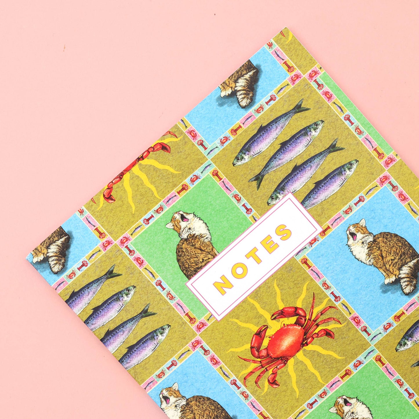 A notebook with a colorful patchwork cover pattern featuring fish, cats, crabs, and a white label reading 'NOTES' in yellow, photographed on a solid pink background.