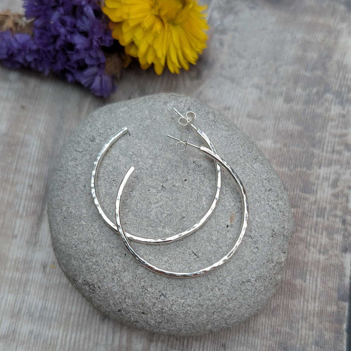 A pair of hammered silver hoop earrings displayed on a smooth gray stone, with dried purple and yellow flowers in the background on a weathered wood surface.