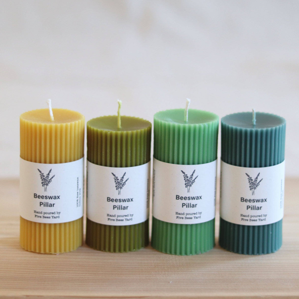 Four beeswax pillar candles in yellow, olive green, mint green, and dark teal, arranged in a row on a light wooden surface with a soft neutral background.