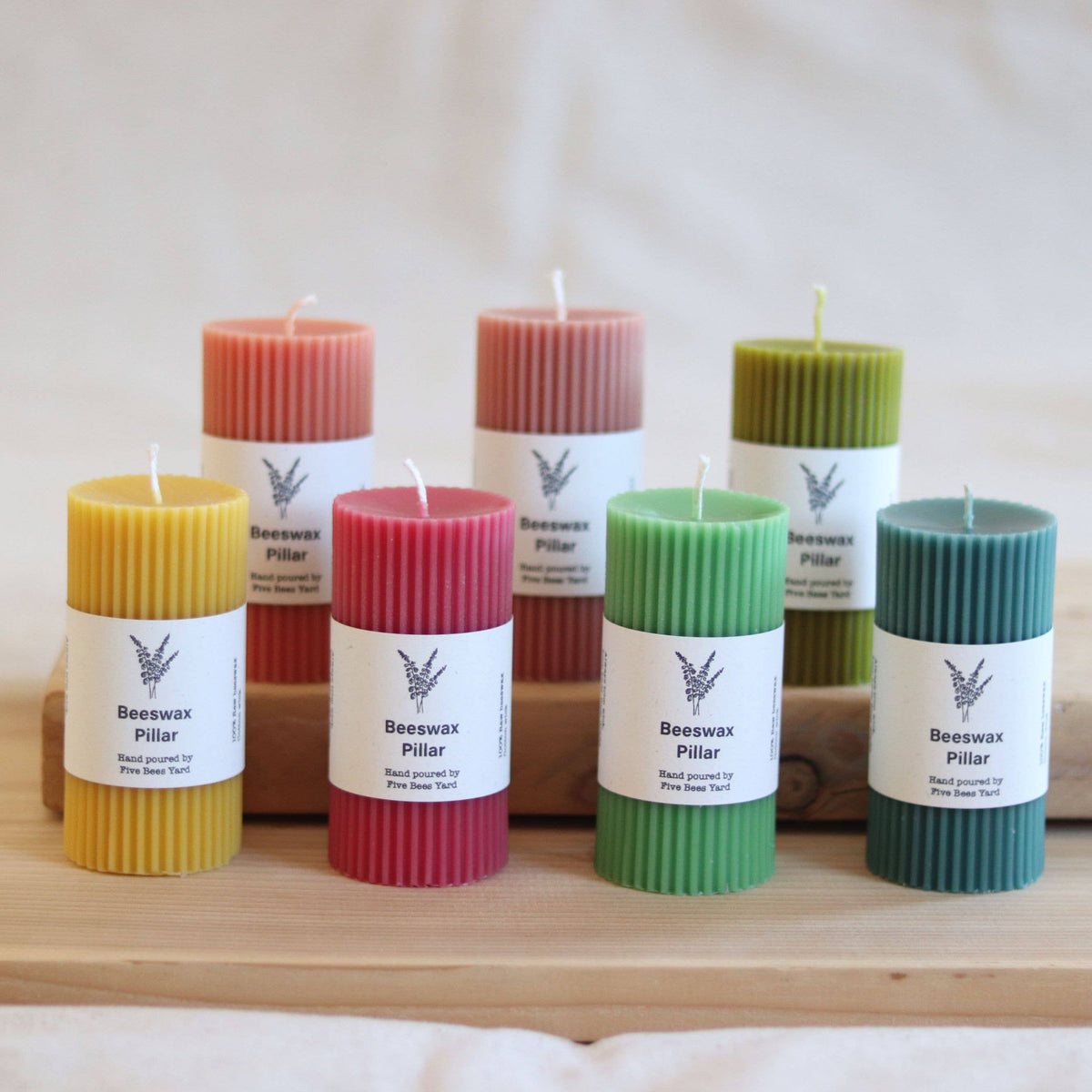 Six ribbed beeswax pillar candles in assorted colors including yellow, coral, pink, green, and teal, arranged on a wooden surface with white paper labels reading 'Beeswax Pillar' and 'Hand poured by Pine Tree Wax Co.'.
