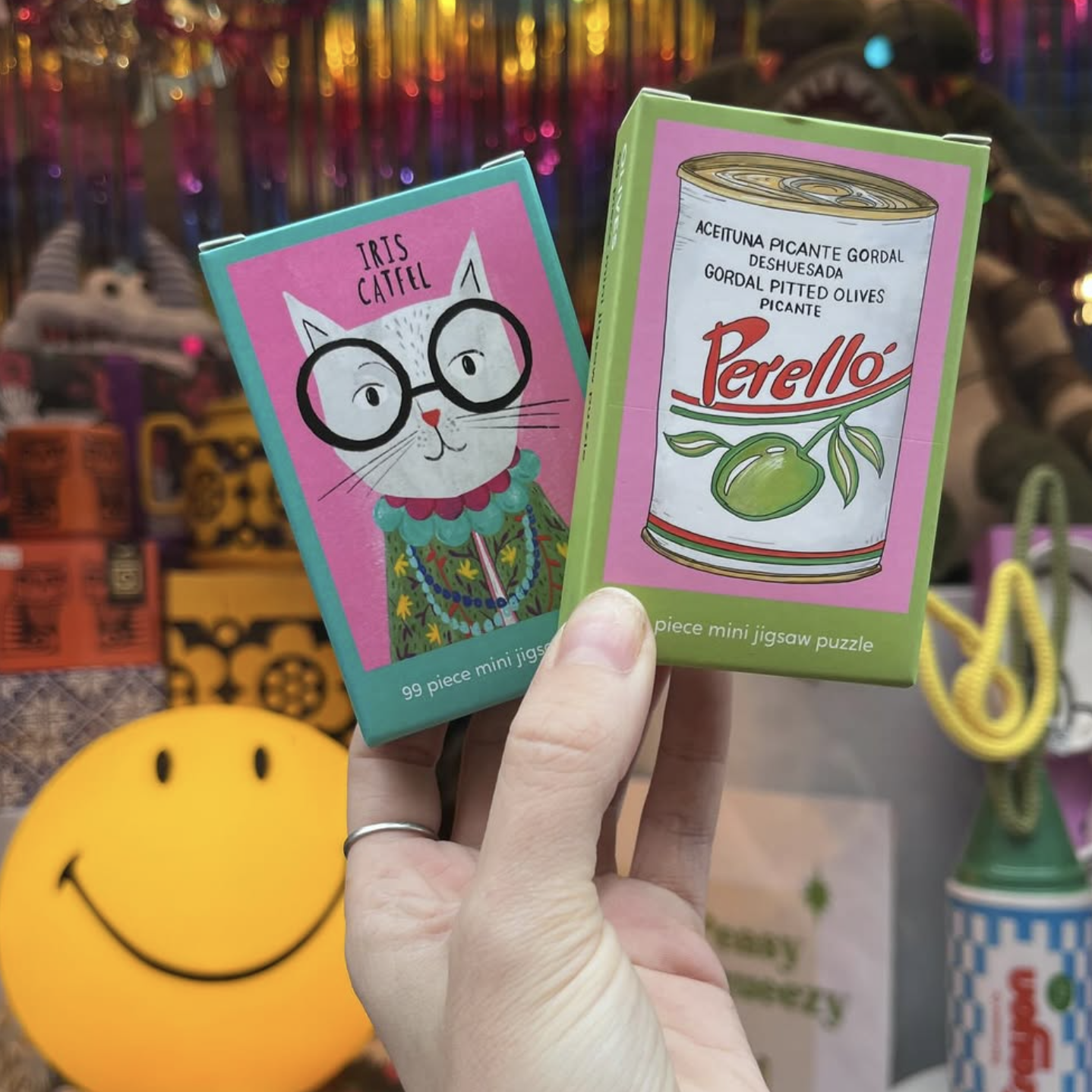 Two mini jigsaw puzzles with a cat and olive oil can design held by a hand against a colorful background.