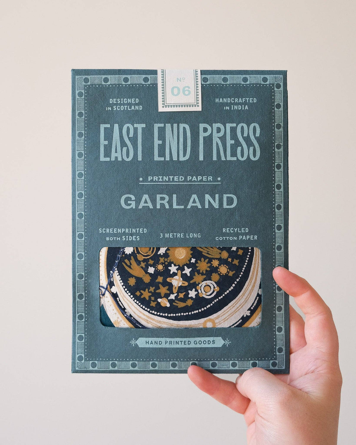 A hand holds a package of East End Press Garland printed paper, model 06. The packaging is a dark teal-green color with light blue-green text and decorative borders, featuring a clear window that shows a stack of patterned circular paper discs inside.
