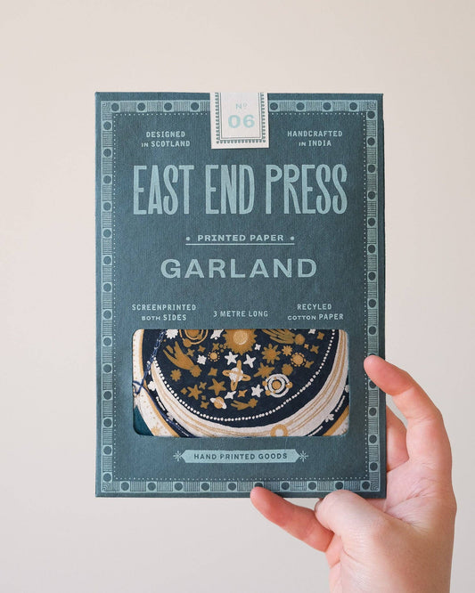 A hand holds a package of East End Press Garland printed paper, model 06. The packaging is a dark teal-green color with light blue-green text and decorative borders, featuring a clear window that shows a stack of patterned circular paper discs inside.