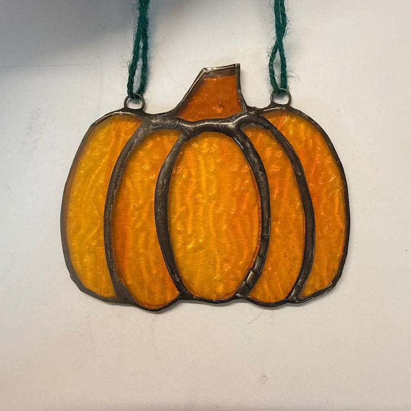 A pumpkin-shaped stained glass ornament with textured orange and yellow glass panels outlined in dark metal, hanging from a green string against a plain off-white background.