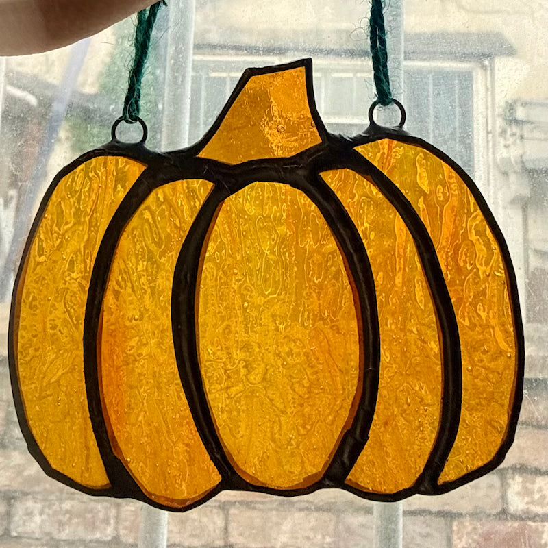 A stained-glass-style pumpkin ornament with textured amber and orange panels outlined in black, hanging by a dark green string in front of a window.