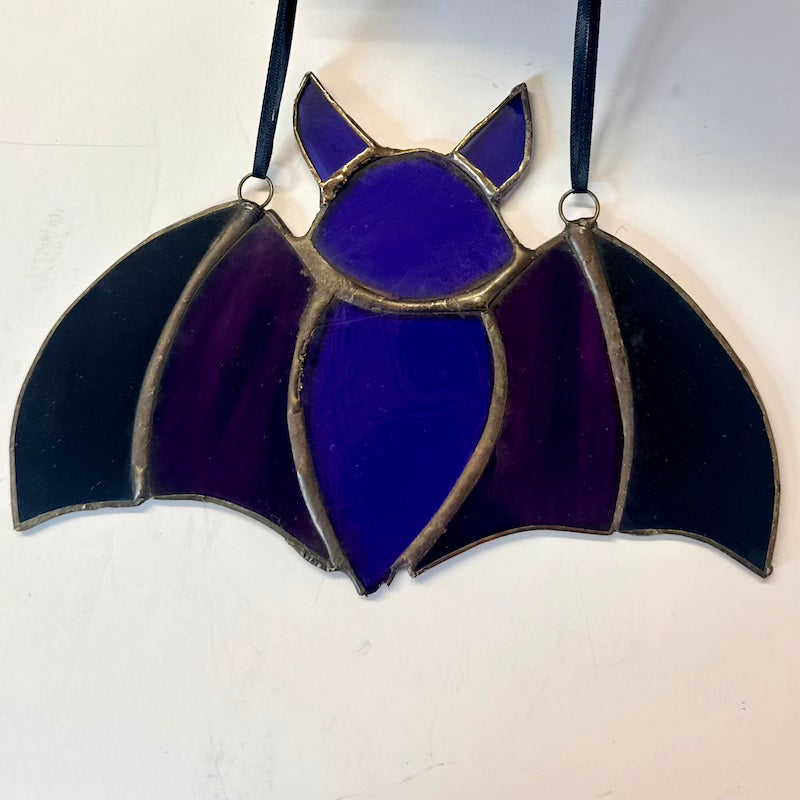 A bat-shaped stained glass hanging ornament with a deep purple body, black wings, and gold-toned metal outlines, suspended by black ribbons against a plain off-white background.