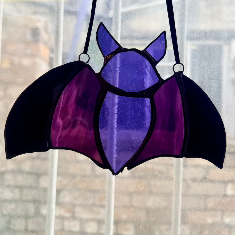 A bat-shaped stained glass hanging ornament with purple and black panels, suspended by two black cords in front of a window.