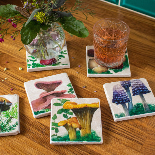 An assortment of six stone coasters with different hand painted woodland mushroom designs with a chanterelle mushroom coaster in the front. On the upper right hand side a drink rests on top of one of the coasters. On the top left there is a round vase of flowers on top of a coaster.