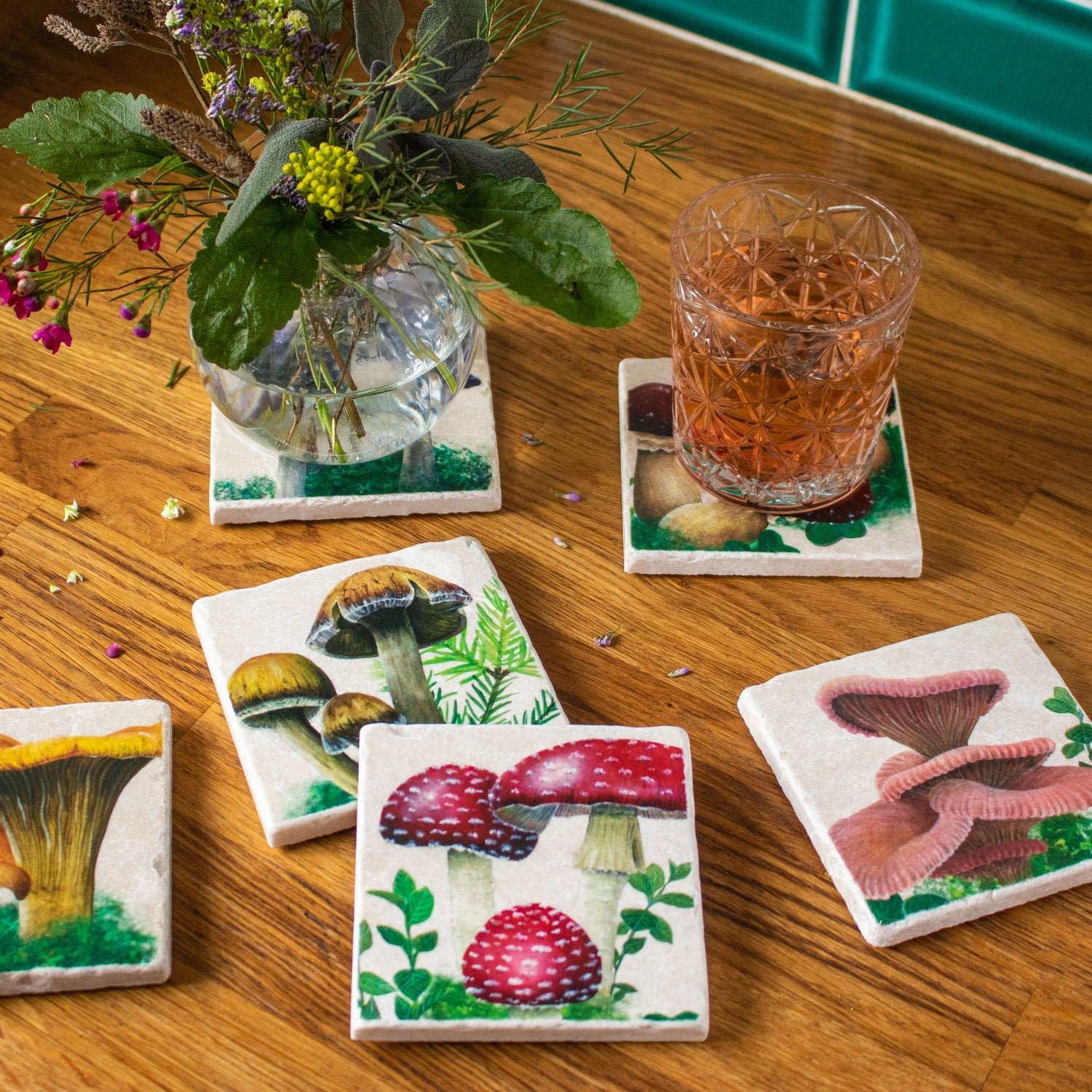 An assortment of six stone coasters with different hand painted woodland mushroom designs with a fly agaric mushroom coaster in front. On the upper right hand side a drink rests on top of one of the coasters. On the top left there is a round vase of flowers on top of a coaster.