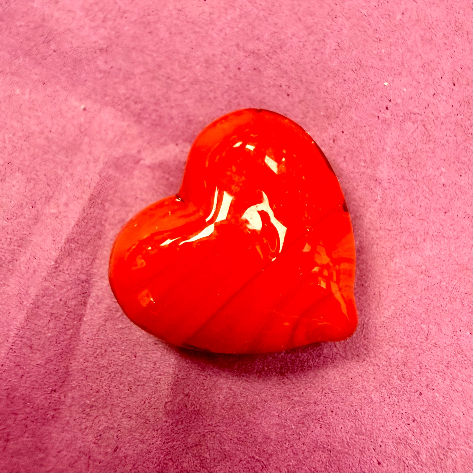A glossy, heart-shaped red wax seal or stamp resting on a textured pink paper background.